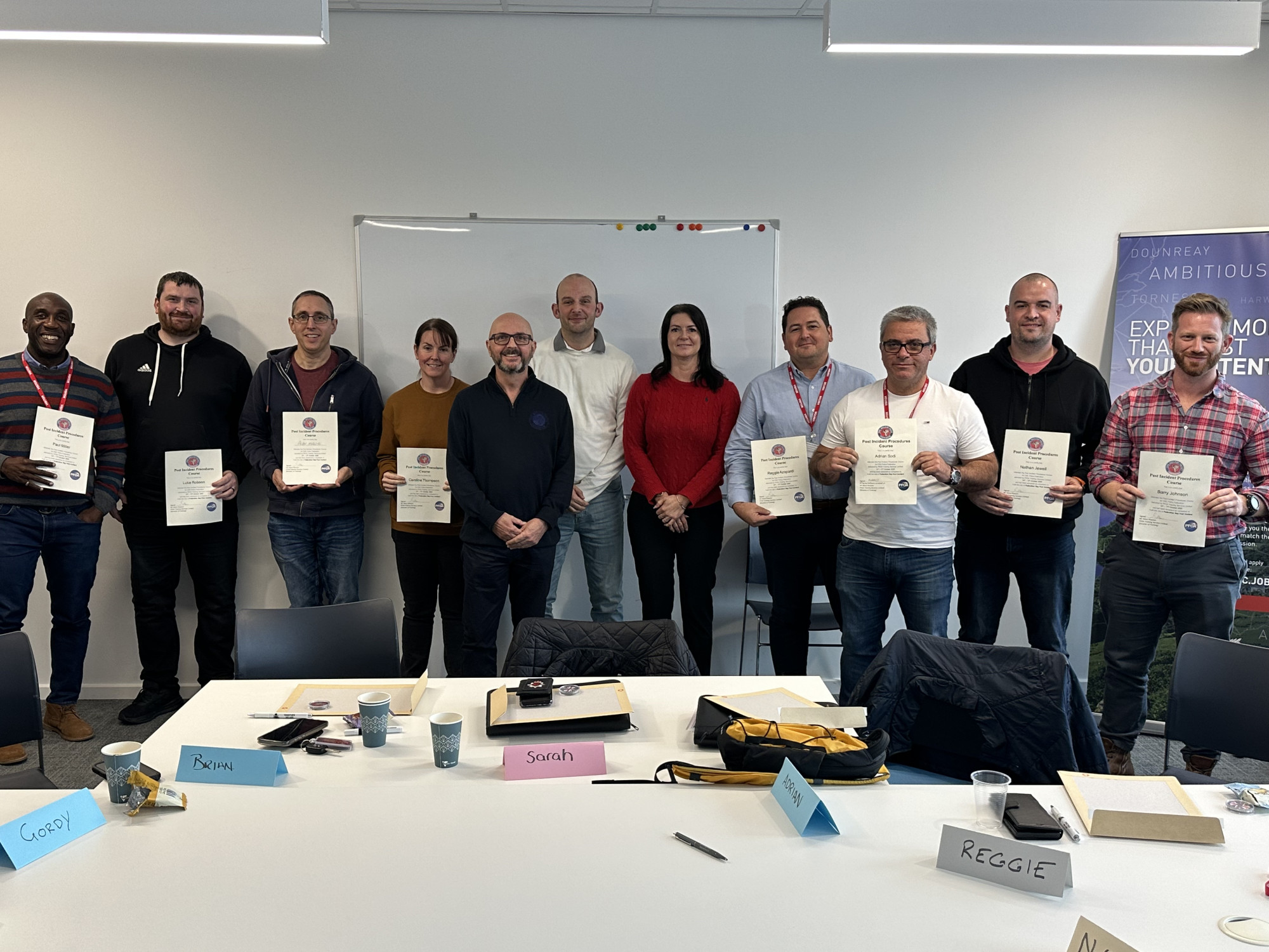 A group of men and women stand in a line facing the camera. Most are holding certificates.