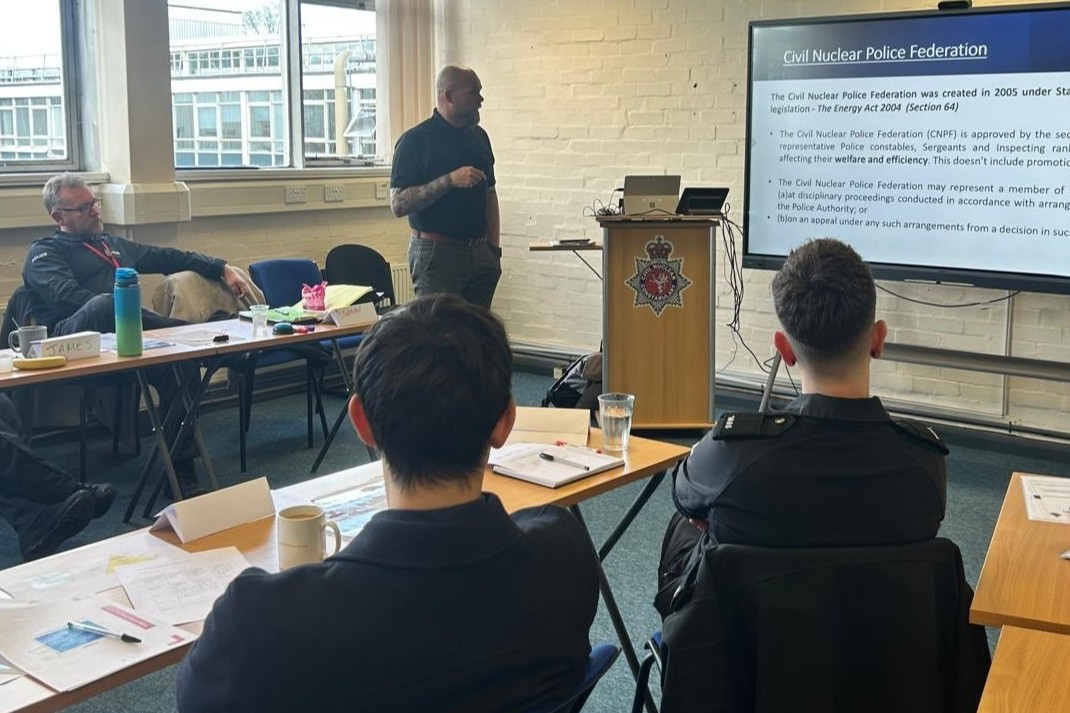 Darren Adams stands at the front of a classroom. Officers are visible watching him as he delivers a presentation.